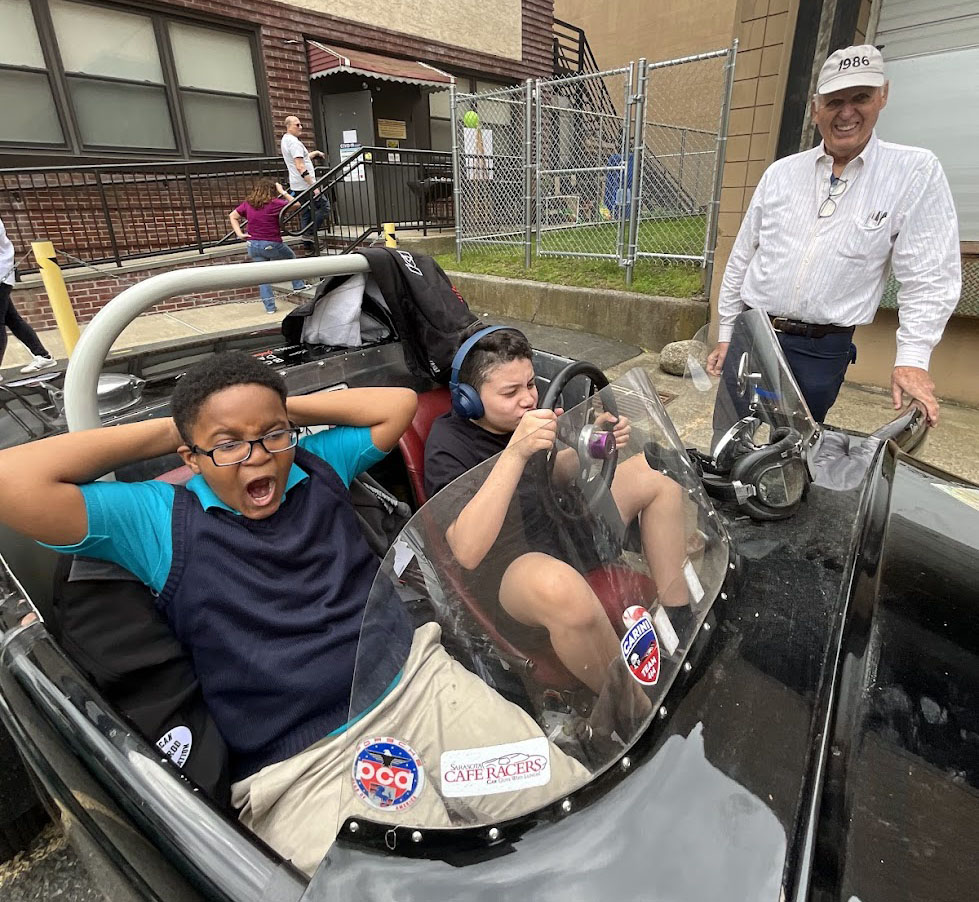 Westbridge studen ts sitting in a race car pretneding to drive, with the owner standing nearby smiling